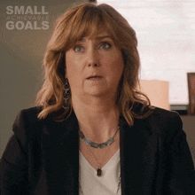a woman wearing a black jacket and a necklace stands in front of a small achievable goals poster