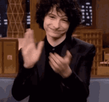 a young man in a suit and black shirt is clapping his hands and smiling .