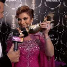 a woman in a pink dress is holding a trophy and a microphone that says trending