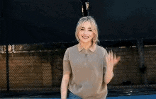 a woman in a polo shirt is waving at the camera while standing in front of a fence .