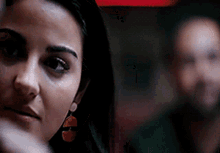 a woman wearing earrings looks at the camera with a man in the background