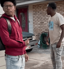 two men are standing in front of a brick building and one is holding a cell phone .