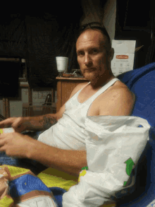 a man in a white tank top sits on a blue couch with a rubbermaid box in the background