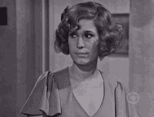 a woman with curly hair is standing in front of a door and looking at the camera in a black and white photo .
