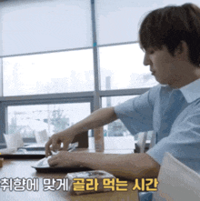 a man sits at a table with a laptop and a foreign language behind him