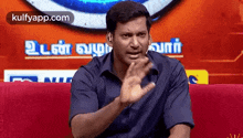 a man in a blue shirt is sitting on a red couch and making a gesture .