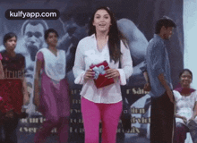 a woman in pink pants and a white shirt is standing in front of a group of people holding a gift box .