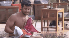 a shirtless man is sitting on a patio holding a towel and a pair of shoes .