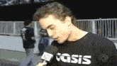 a man wearing a black oasis shirt talks into a microphone