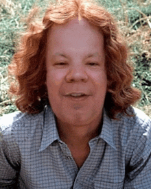 a man with red hair is wearing a plaid shirt and smiling with a clock in the background