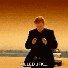a man in a suit is standing on a beach with a car in the background and says `` killed jfk ... '' .
