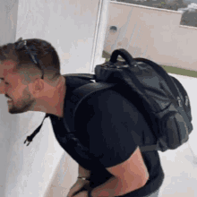 a man wearing a black shirt and a black backpack stands in front of a white wall