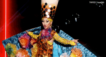 a drag queen is wearing a colorful costume and a bubble tea cup on her head .