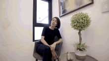 a woman sits in a chair next to a potted plant and a window