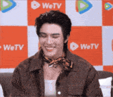 a man wearing a brown jacket and a scarf is smiling in front of a wetv logo