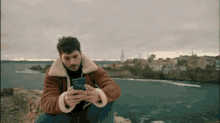 a man in a brown jacket is sitting on a rock looking at his phone