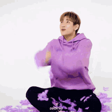 a man wearing a purple hoodie is sitting on the floor with purple feathers around him .