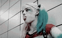 a woman with blue hair is sticking her tongue out while standing in front of a tiled wall .