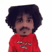 a man with curly hair and a mustache is wearing a red sweatshirt with arabic writing on it .
