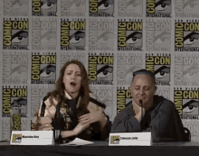a man and a woman are sitting at a table in front of a comic con wall