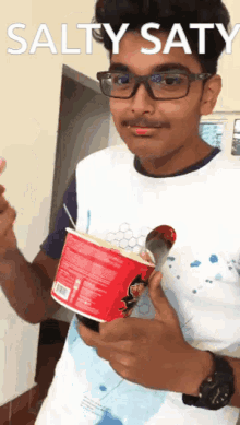 a young man with glasses is holding a cup of noodles with a spoon and the words salty saty above him