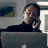 a man in a black turtleneck is talking on a cell phone in front of an apple laptop