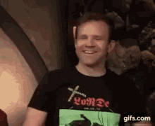 a man wearing a black t-shirt is smiling while standing in front of a rock wall .