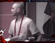 a bald man wearing a white shirt and red beads is sitting at a table