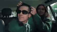 a man wearing sunglasses is sitting in a car with a woman adjusting his hoodie .