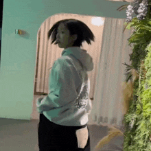 a woman in a white hoodie is standing in front of a green wall with flowers .