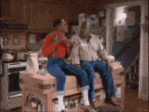 two men are sitting on a wooden counter in a kitchen talking to each other .