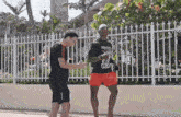 two men are standing next to each other on a sidewalk in front of a white fence .