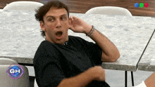 a man is sitting at a table with his mouth open and a gh logo in the corner