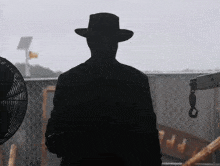 a silhouette of a man wearing a hat and a coat is standing in front of a fan .