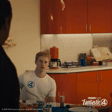 a man is sitting at a table in a kitchen wearing a fantastic 4 shirt
