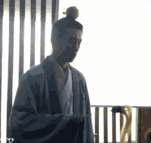 a man with a bun on his head is standing in front of a window holding a sword .