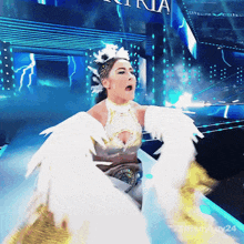 a woman in a white dress with wings is on a stage with a tiffany luv24 logo in the corner