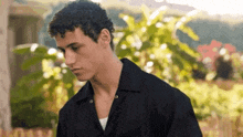 a young man with curly hair is wearing a black shirt and standing in front of a bush .