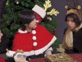 a man in a santa cape sits next to a christmas tree
