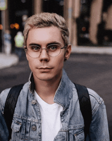 a young man wearing glasses and a denim jacket with a backpack