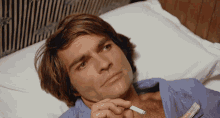 a man in a blue shirt is laying on a bed holding a cigarette