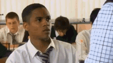 a man in a white shirt and tie is sitting at a desk in a classroom with other students .