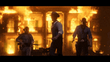 three men in cowboy hats stand in front of a burning building