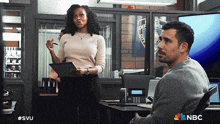 a man sits at a desk while a woman stands behind him with a clipboard