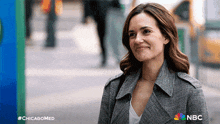 a woman in a gray coat is smiling in front of a nbc sign