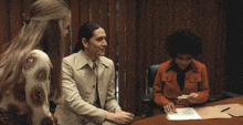 a woman in a red jacket sits at a desk talking to two other women