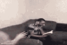 a man is sitting on a couch reading a book while a woman throws a frisbee at him .