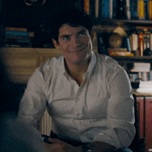 a man in a white shirt is sitting in front of a bookshelf and smiling