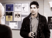 a man in a black jacket is standing in front of a bulletin board holding a cup .