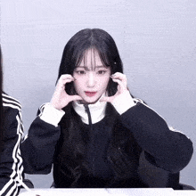 a girl wearing a black jacket with white stripes on the sleeves is making a heart shape with her hands